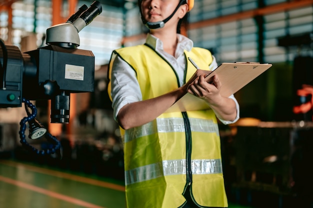 Industria dell'ingegnere donna asiatica che indossa l'elmetto protettivo e tiene la lista di controllo in piedi nell'area della macchina