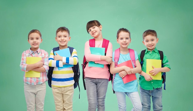 infanzia, educazione prescolare, apprendimento e concetto di persone - gruppo di bambini sorridenti felici con zaini e quaderni su sfondo verde lavagna