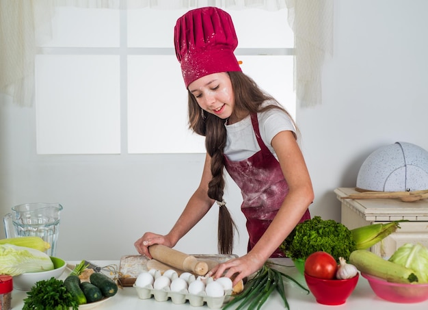 Infanzia felice bambino felice indossare cuoco uniforme chef ragazza con cappello e grembiule bambino cucinare cibo in cucina scegliendo un piccolo aiutante di carriera rotolare la pasta con il mattarello culinario e cucina