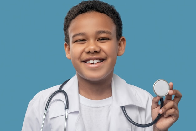 Infanzia, gioia. Felice ragazzo ridente dalla pelle scura in camice bianco che mostra uno stetoscopio medico su sfondo blu in studio