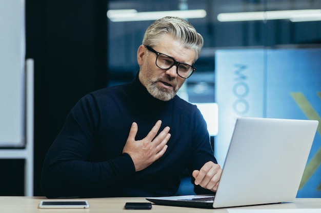 Infarto dell'uomo d'affari sul posto di lavoro uomo maturo dai capelli grigi che lavora al computer portatile ha un forte dolore toracico