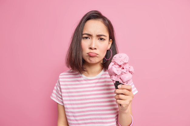 Infelice scontento La donna asiatica sembra offesa tiene un grande cono gelato continua a dieta ha la tentazione di mangiare cibo spazzatura ha i capelli corti scuri vestiti con una maglietta a righe casual isolata sul muro rosa