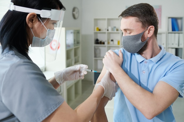 Infermiera asiatica in abbigliamento da lavoro uniforme e protettivo che fa iniezione al giovane durante la vaccinazione contro il covid nelle cliniche contemporanee