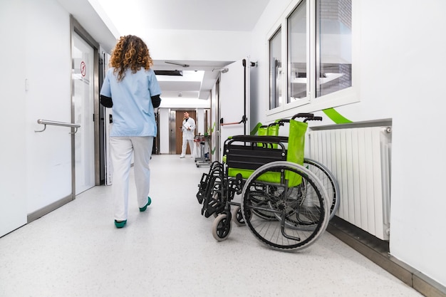 Infermiera che si allontana in un luminoso corridoio dell'ospedale con una sedia a rotelle verde vuota sul lato
