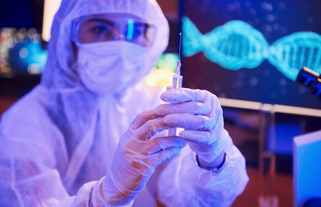 Infermiera in maschera e uniforme bianca, con in mano una siringa e seduta in laboratorio illuminato al neon con computer e apparecchiature mediche alla ricerca del vaccino contro il coronavirus