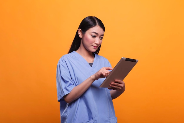 Infermiera ospedaliera abbastanza asiatica che indossa un'uniforme medica blu mentre utilizza un tablet moderno su sfondo arancione. Membro del personale sanitario della clinica che utilizza tablet digitale. Riprese in studio.