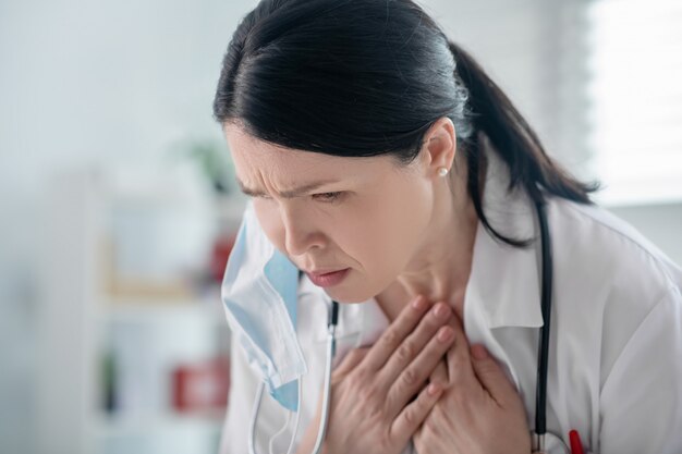 Infezione da virus. Brutta donna adulta dai capelli scuri in camice bianco e uno stetoscopio sul collo tenendosi per mano per la gola