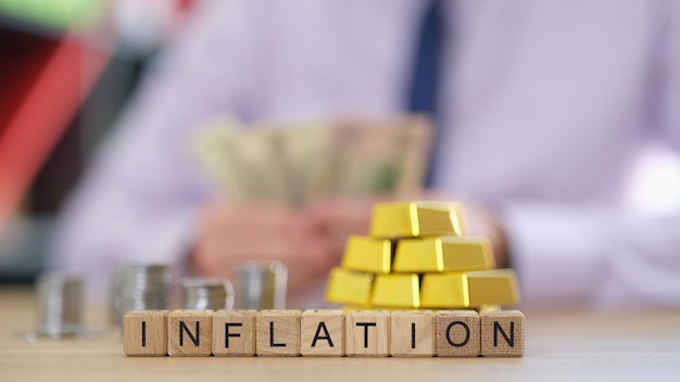 Inflazione di lettere su cubi di legno con lingotti d'oro e pile di monete sul tavolo uomo d'affari sfocato