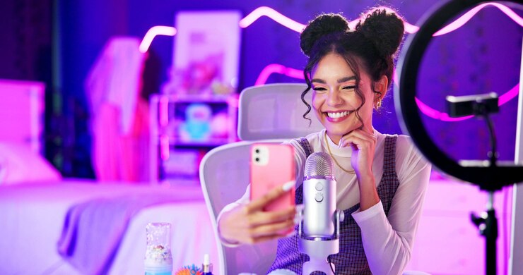 Foto influencer selfie e donna con aggiornamento in streaming dal vivo in camera da letto al neon con fan o seguaci del pubblico dei social media creatore di contenuti eccitato giocatore o streamer tramite microfono smartphone e gioco a casa