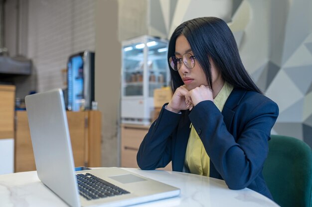 Informazioni importanti. Piuttosto giovane donna dai capelli scuri con gli occhiali che si toccano le mani al mento guardando attentamente il laptop seduto a lavorare in ufficio