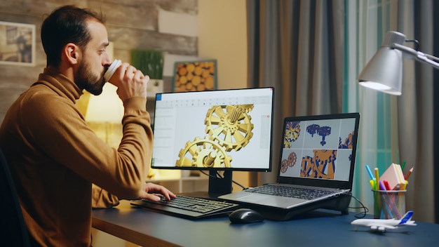 Ingegnere barbuto che beve un sorso di caffè mentre lavora al computer. Produzione di ingranaggi.