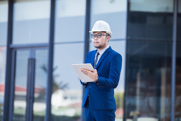 Ingegnere bello che indossa un vestito e un casco o un casco bianco sullo sfondo dell'edificio Ingegnere che usa un tablet per controllare la costruzione di edifici moderni spazio di copia per il testo