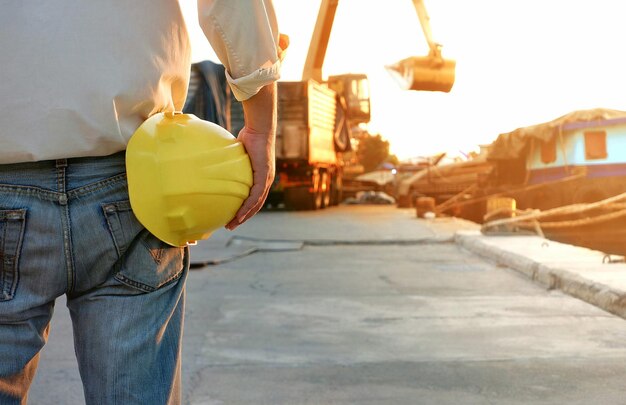 Ingegnere che tiene il casco giallo e guarda l'escavatore a cucchiaia rovescia che trasferisce la chiatta di forma di sabbia al porto