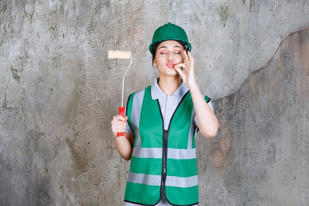 Ingegnere donna con casco verde che tiene in mano un rullo di rifinitura per la pittura murale e mostra un segno positivo con la mano