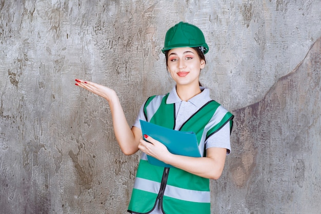 Ingegnere donna con casco verde che tiene una cartella blu e indica qualcuno intorno