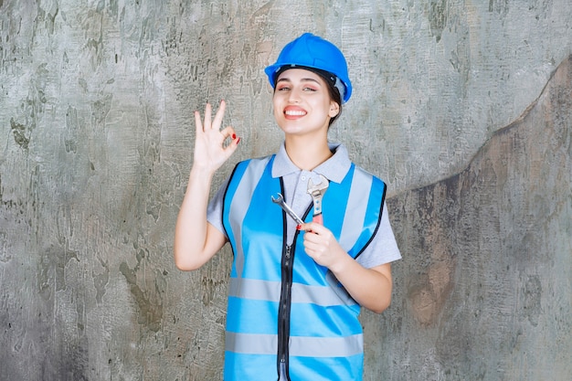 Ingegnere donna in uniforme blu e casco che tiene in mano una chiave metallica e mostra un segno positivo con la mano