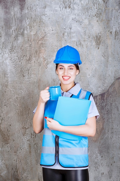 Ingegnere donna in uniforme blu e casco con in mano una tazza da tè blu e una cartella di rapporto blu