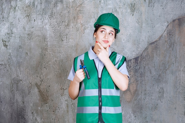 Ingegnere femminile in casco verde che tiene le pinze per un lavoro di riparazione e sembra confuso e premuroso.