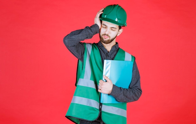 Ingegnere in uniforme verde e casco in possesso di una cartella blu, sembra stanco e assonnato.