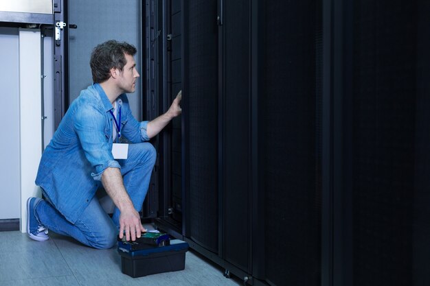 Ingegnere maschio bello bello seduto su un ginocchio e prendendo il server rack mentre lo installa nel server di rete