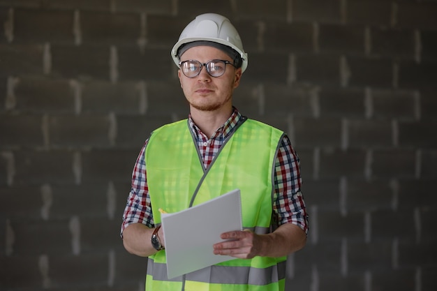 Ingegnere maschio europeo in casco bianco e giubbotto di sicurezza nel cantiere che studia il