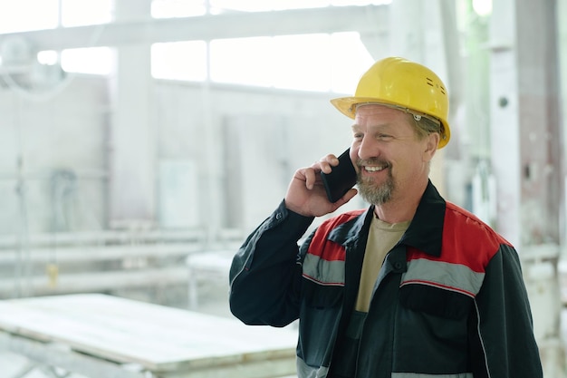 Ingegnere sorridente maturo che ha conversazione sul telefono cellulare mentre lavora in fabbrica