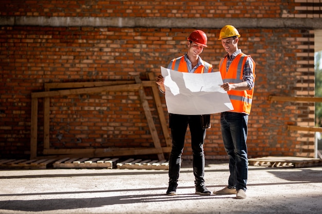 Ingegnere strutturale e architetto vestito con giubbotti e caschi da lavoro arancioni discutono la documentazione su uno sfondo di muro di mattoni.