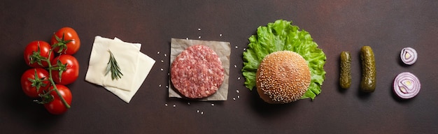 Ingredienti dell'hamburger cotoletta cruda, pomodori, lattuga, panino, formaggio, cetrioli e cipolla su fondo arrugginito. Vista dall'alto