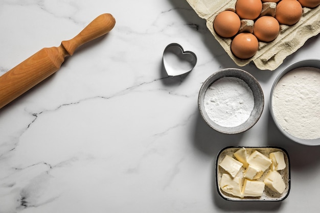 Ingredienti e utensili per la cottura di biscotti di pasta frolla su fondo di marmo Uova farina burro zucchero a velo a forma di cuore tagliabiscotti mattarello in legno