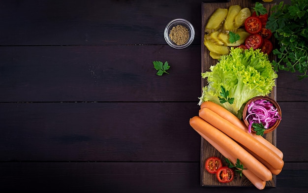 Ingredienti per hot dog con salsiccia cetriolo pomodori cipolle rosse e lattuga su sfondo di legno scuro hot dog estivo vista superiore