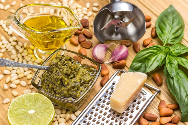 Ingredienti per il pesto fatto in casa. Parmigiano su grattugia. pesto in tazza di vetro. Limone e foglie, basilico e olio. Aglio e Pinoli. Trituratore di metalli. Vista dall'alto. Fondo in legno chiaro.