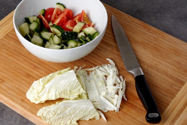 Ingredienti per insalata. Ciotola di pomodori e cetrioli tritati e un tagliere con cavolo cinese tritato e un coltello su un tavolo scuro.