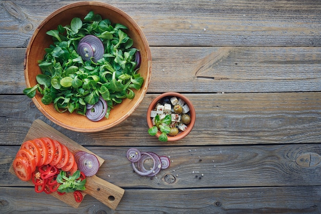 Ingredienti per insalata di verdure su una superficie di legno: foglie di lattuga, pomodori, peperoncino, cipolle, olive, olio e formaggio