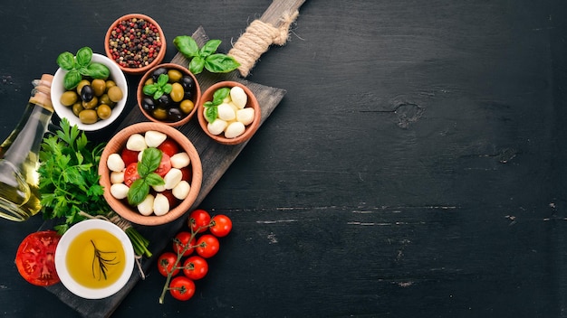 Ingredienti per l'insalata caprese italiana Mozzarella pomodorini basilico foglie olive olio pepe Su fondo di legno nero Spazio libero per il testo