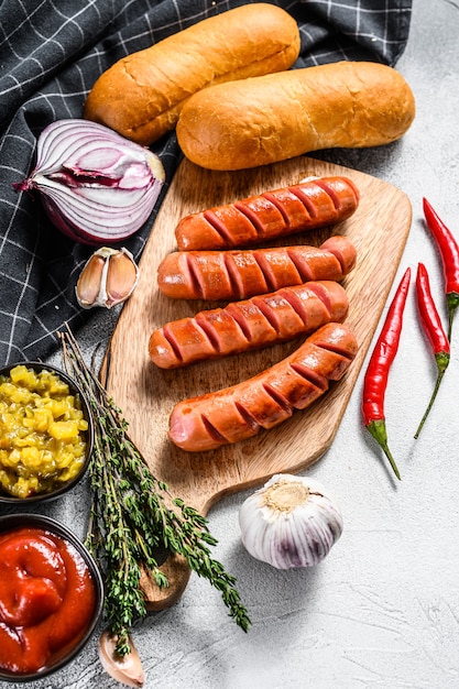 Ingredienti per preparare hot dog fatti in casa. Salsicce, panini appena sfornati, senape, ketchup, cetrioli. Sfondo bianco. Vista dall'alto