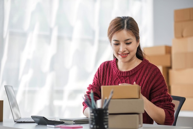 Iniziando una piccola impresa imprenditrice di donna asiatica indipendente sorridendo usando un computer portatile con un allegro successo di articoli di confezione di marketing online e concetto di consegna di PMI
