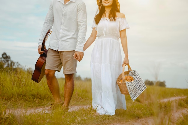 Innamorati felici camminano e si tengono per mano sul prato con tenerezza per trovare un posto dove rilassarsi. Un uomo con una chitarra e una donna con una scatola da picnic.