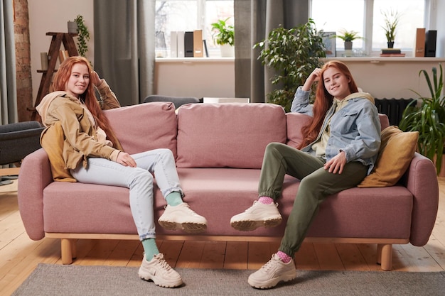 Inquadratura ampia di due belle giovani sorelle gemelle caucasiche con lunghi capelli rossi che indossano abiti casual seduti rilassati sul divano in soggiorno