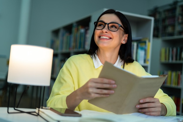 Inquadratura dal basso del libro di lettura della giovane donna in biblioteca e sorridente signora bruna con i denti che studia al liceo Foto d'archivio