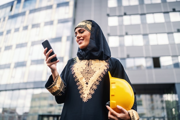 Inquadratura dal basso di attraente donna musulmana sorridente in piedi davanti all'edificio aziendale, utilizzando smart phone e tenendo il casco sotto l'ascella. Anche le donne possono essere grandi architetti.