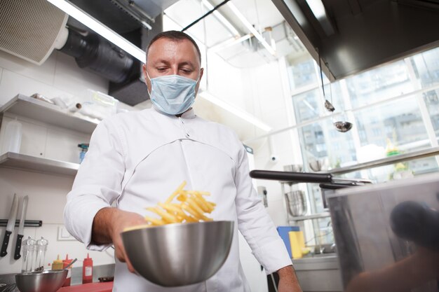 Inquadratura dal basso di uno chef che indossa una maschera medica, che prepara patatine fritte al ristorante