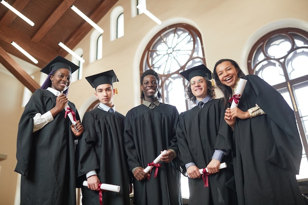 Inquadratura dal basso in un gruppo multietnico di giovani che indossano abiti da laurea e guardano la telecamera sorridendo felicemente mentre si trovano nell'auditorium della scuola