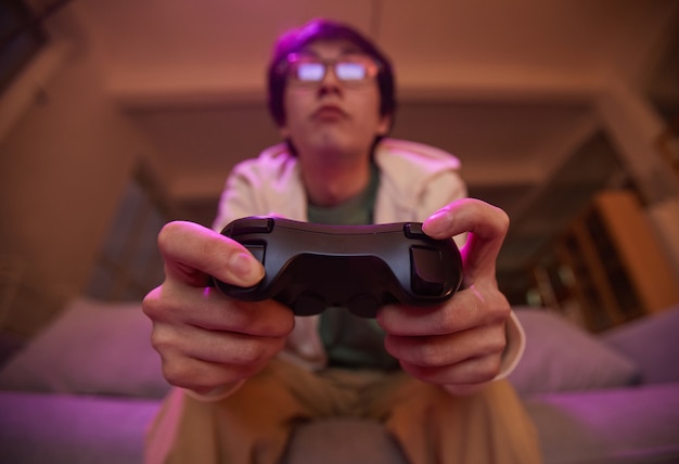 Inquadratura dal basso su un giovane asiatico che tiene in mano il gamepad e gioca ai videogiochi a casa illuminata da luce viola, copia spazio