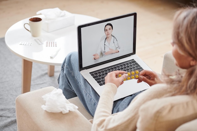 Inquadratura dall'alto di una donna seduta sul divano che sta a casa a parlare con il medico durante una videochiamata utilizzando il laptop