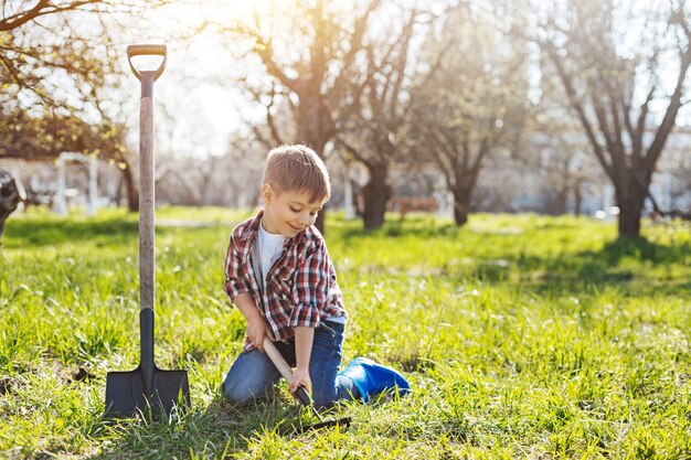 Inquadratura di un bambino seduto su un prato verde primaverile e scavando una buca nel terreno per futuri alberi da frutto in primavera