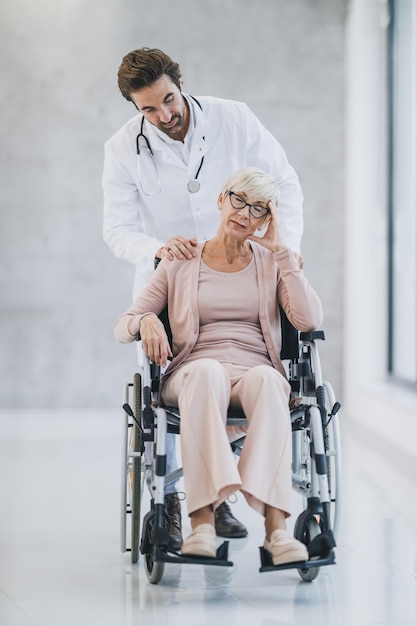 Inquadratura di un giovane medico che spinge la sua paziente anziana in sedia a rotelle e parla con lei.