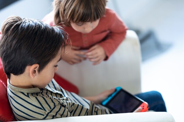 Inquadratura di un ragazzino che gioca ai videogiochi mentre è seduto sul divano e suo fratello lo guarda a casa.
