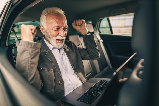 Inquadratura di un uomo anziano allegro che usa il laptop e celebra il lavoro di successo mentre è seduto sul sedile posteriore di un'auto durante i suoi spostamenti di lavoro.