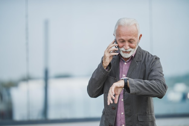 Inquadratura di un uomo d'affari senior di successo che parla su uno smartphone mentre guarda l'orologio di fronte a un edificio aziendale.