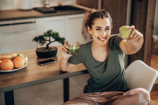 Inquadratura di una donna sorridente con in mano un bicchiere di frullato e mela verde a casa al mattino. Guardando la fotocamera.